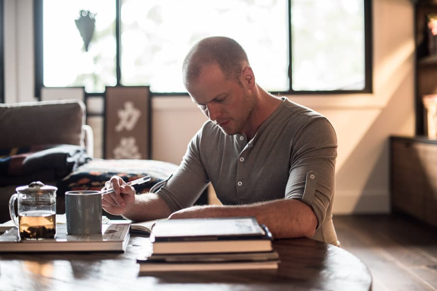 Picture of Tim Ferriss seated on the floor in a living room. Tim is reading and taking notes with a cup of tea in front of him. Tim is Caucasian with a buzzed haircut, stubble, and wearing a henley.