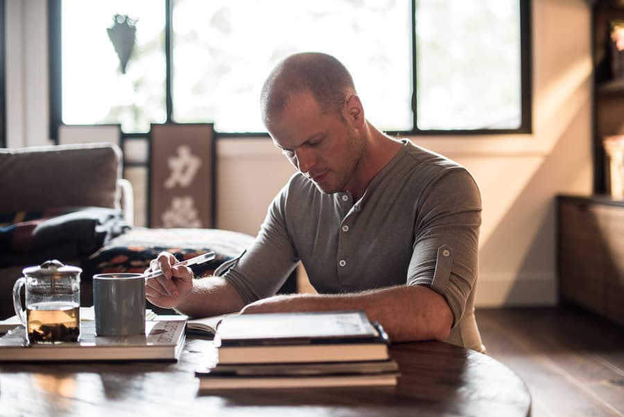 Picture of Tim Ferriss seated on the floor in a living room. Tim is reading and taking notes with a cup of tea in front of him. Tim is Caucasian with a buzzed haircut, stubble, and wearing a henley.